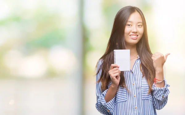 年轻的亚洲妇女拿着笔记本在孤立的背景下指向和显示与拇指到一边 快乐的脸微笑 — 图库照片