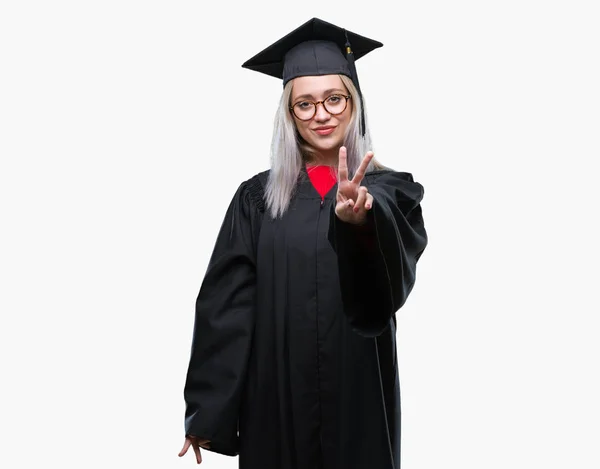 勝利のサインを行うカメラでウィンクしている幸せそうな顔を浮かべて孤立の背景の上の大学院の制服を着て若いブロンドの女性 — ストック写真