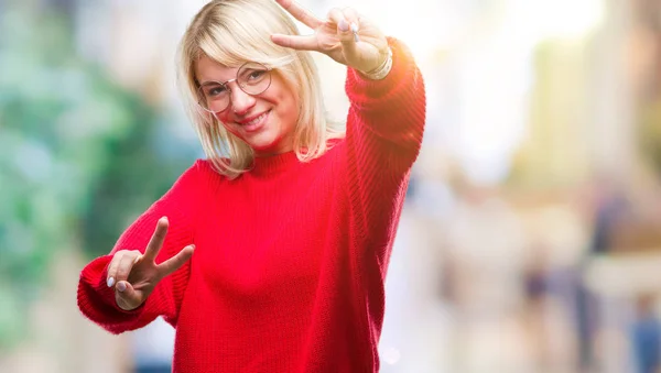 若い美しい金髪女性の勝利のサインをやって指を表示するカメラを笑みを浮かべて隔離された背景にセーターとメガネを着用します — ストック写真