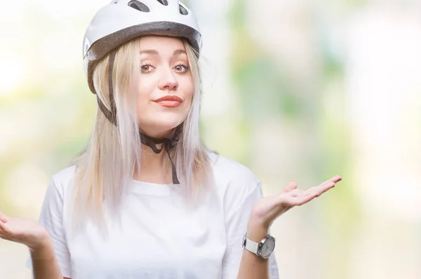 年轻的金发碧眼的女人戴着骑自行车的安全帽 在孤立的背景无线索和混乱的表情与举起的胳膊和手 怀疑概念 — 图库照片