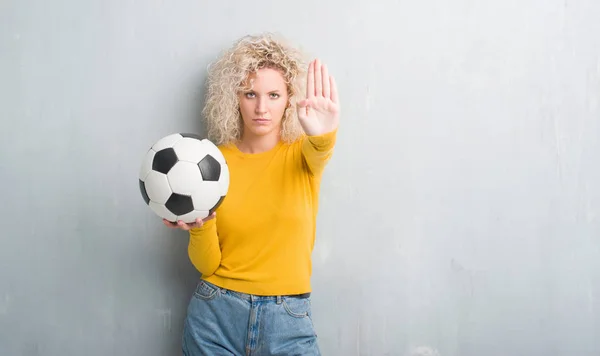 Young blonde woman over grunge grey background holding soccer football ball with open hand doing stop sign with serious and confident expression, defense gesture