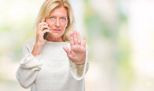 Middle age blonde woman talking on smartphone over isolated background with open hand doing stop sign with serious and confident expression, defense gesture