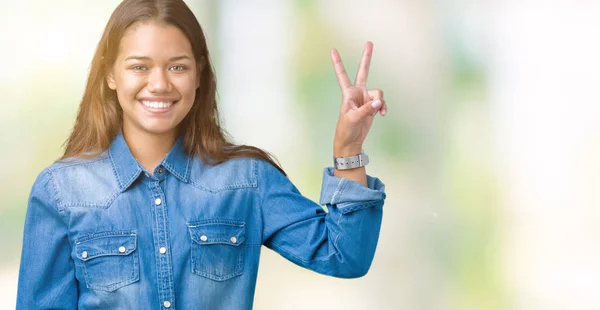 Young beautiful brunette woman wearing blue denim shirt over isolated background smiling with happy face winking at the camera doing victory sign. Number two.