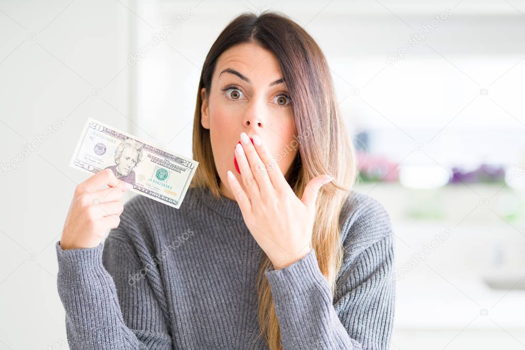 Young beautiful woman holding 20 dollars bank note at home cover mouth with hand shocked with shame for mistake, expression of fear, scared in silence, secret concept
