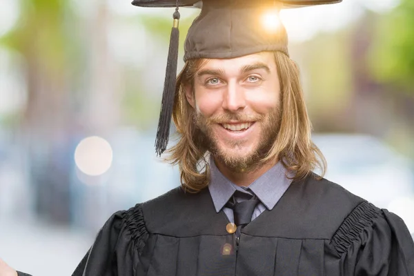 Young handsome graduated man with long hair over isolated background amazed and smiling to the camera while presenting with hand and pointing with finger.