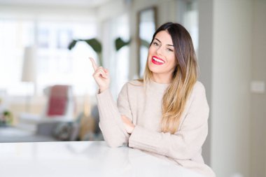 Kameraya bakarak yan el ve parmak ile işaret yüzünde, evde kocaman bir gülümseme ile kış kazak giyen genç güzel kadın.