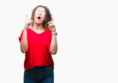 Güzel esmer kıvırcık saçlı Genç kız izole arka plan üzerinde gözlük hayret ve yukarıya bakıp parmakları ile işaret şaşırttı ve silah kaldırdı.