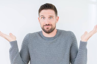 Üzerinde rahat kazak giyen genç yakışıklı adam arka plan clueless ve şaşkın bir ifadeyle kollar ve eller kaldırdı izole. Şüphe kavramı.