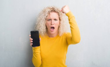 Boş ekran smartphone rahatsız ve öfke, çılgın ve yükseltilmiş el, öfke kavramı ile diye bağıran ile sinirli bağırarak gösterilen grunge gri arka plan üzerinde sarışın genç kadın