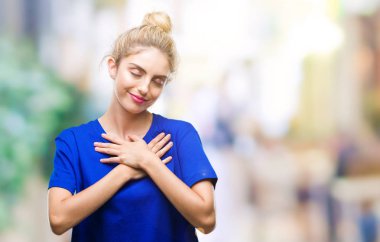 Göğüs kapalı gözlerle ellerinde ve yüzünde minnettar jest ile gülümseyen izole arka plan üzerinde mavi t-shirt giyen genç güzel sarışın ve mavi gözlü kadın. Sağlık kavramı.