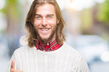 Genç yakışıklı adam uzun saçlı hareketi el ile mutlu yaşasın yapıyor izole arka plan üzerinde kış kazak giyiyor. Kameraya bakarak başarı gösteren ile ifade onaylama.