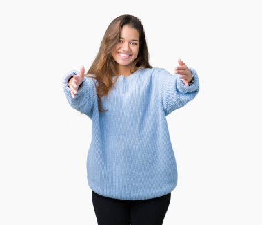 Kamera ile gülümseyen bakarak izole arka plan üzerinde mavi kış kazak giyen genç güzel esmer kadın kolları sarılmak için açın. Neşeli ifade kucaklayan mutluluk.