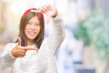 Genç Asyalı kadın elle yapma çerçeve ve parmakları ile mutlu yüz gülümseyen izole arka plan üzerinde kış kazak giyiyor. Yaratıcılık ve Fotoğrafçılık kavramı.