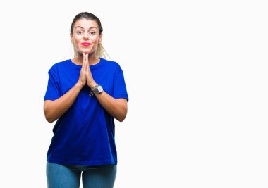 Ellerle birlikte AF emin gülümseyen için soran dua izole arka plan üzerinde rahat mavi t-shirt giyen genç güzel kadın.