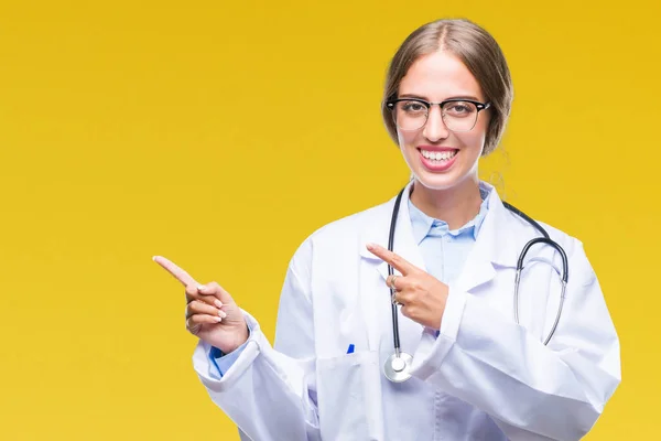 美丽的年轻金发碧眼的医生妇女穿着医疗制服在孤立的背景微笑着 看着相机指向用两只手和手指的一侧 — 图库照片