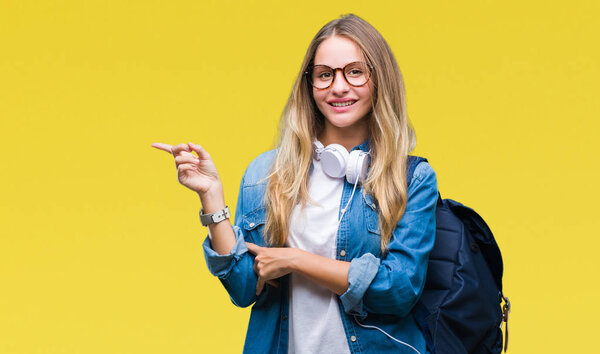 Young beautiful blonde student woman wearing headphones and glasses over isolated background with a big smile on face, pointing with hand and finger to the side looking at the camera.