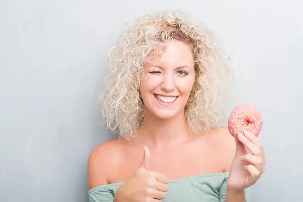 グランジに若いブロンドの女性灰色背景笑顔の サイン 優秀な兆候を親指をやって幸せピンクのドーナツを食べる — ストック写真