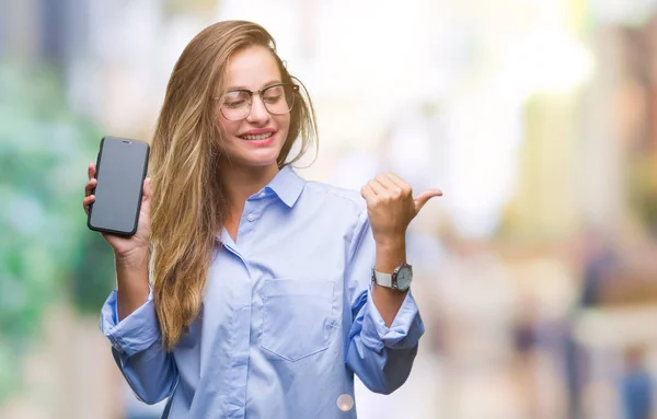 Young beautiful blonde business woman showing screen of smartphone over isolated background pointing and showing with thumb up to the side with happy face smiling