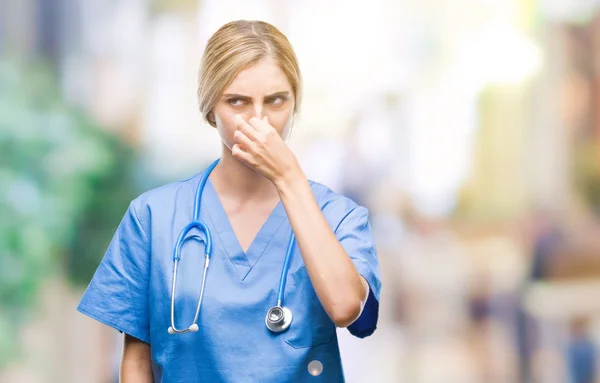 Young beautiful blonde doctor surgeon nurse woman over isolated background smelling something stinky and disgusting, intolerable smell, holding breath with fingers on nose. Bad smells concept.