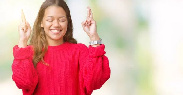 年轻美丽的黑发女人穿着红色的冬季毛衣在孤立的背景微笑交叉手指与希望和眼睛闭上 运气和迷信的概念 — 图库照片