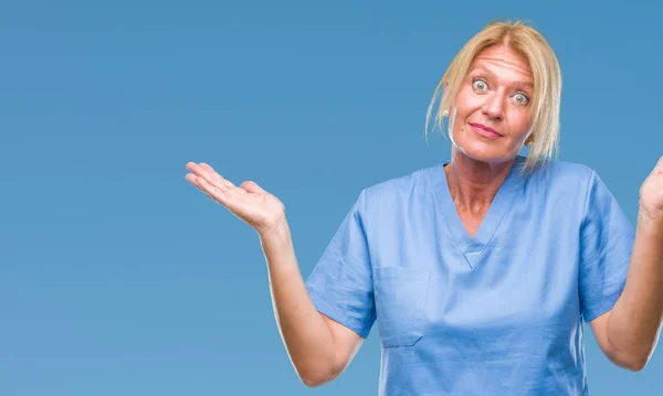 中年金发碧眼的妇女穿着医生护士制服在孤立的背景无线索和混乱的表情与手臂和手抬起来 怀疑概念 — 图库照片