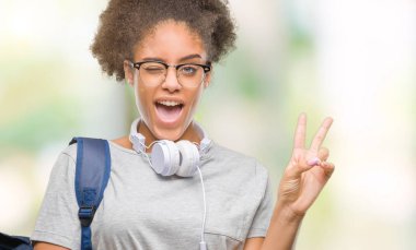Mutlu yüz zafer işareti yaparken kameraya göz kırpıyor ile gülümseyen izole arka plan üzerinde kulaklık ve sırt çantası giyen genç afro Amerikan öğrenci kadın. İki numaralı.