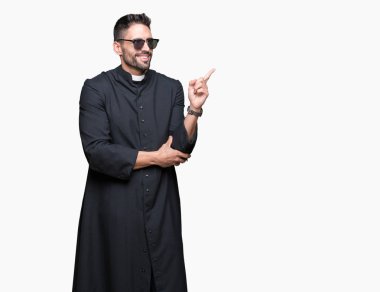 Genç Hıristiyan giyen güneş gözlüğü kameraya bakarak yan el ve parmak ile işaret yüzünde kocaman bir gülümseme ile izole arka plan üzerinde rahip.