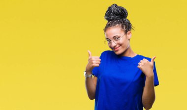 El ile pozitif jest yapıyor izole arka plan başarı işareti üzerinde gözlük takan genç örgülü saç Afro-Amerikan Kız başparmak gülümseyen ve mutlu. Neşeli bir ifadeyle, kazanan jest kameraya bakıyor.