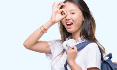 Sırt çantası ve kulaklık ile mutlu yüz el parmaklarının arasından seyir göz üzerinde ok işaretiyle yapıyor gülümseyen izole arka plan üzerinde giyen genç Asyalı kadın