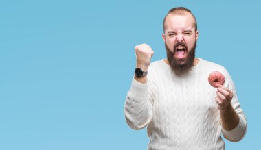 Genç beyaz hipster adam tatlı çörek üzerinde izole yeme arka plan ile öfke, çılgın ve yükseltilmiş eliyle diye bağıran rahatsız ve hayal kırıklığına uğramış öfke kavramı bağırarak