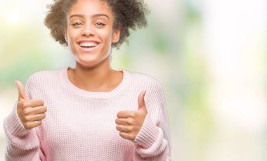 El ile pozitif jest yapıyor izole arka plan başarı işareti üzerinde kış kazak giyen genç afro Amerikan kadın başparmak gülümseyen ve mutlu. Neşeli bir ifadeyle, kazanan jest kameraya bakıyor.