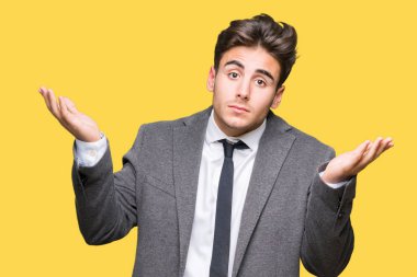 Üzerinde takım elbise ve kravat giyen genç iş adamı arka plan clueless ve şaşkın bir ifadeyle kollar ve eller kaldırdı izole. Şüphe kavramı.