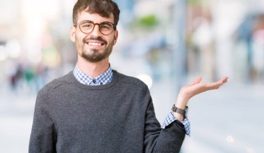 Genç yakışıklı zeki adam neşeli sunulması ve kameraya bakarak avucunun ile işaret gülümseyen izole arka plan üzerinde gözlük.