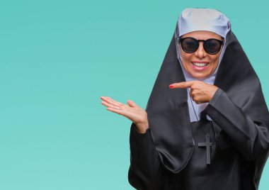 Orta yaş üst düzey Katolik Rahibe kadın hayrete ve el ile sunulması ve parmak ile işaret ederken kameraya gülümseyen izole arka plan üzerinde güneş gözlüğü takıyor.