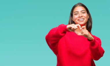 Kalp simgesi ve elleriyle şekil gösterilen aşık gülümseyen izole arka plan üzerinde kış kazak giyen genç güzel Arap kadın. Romantik kavramı.