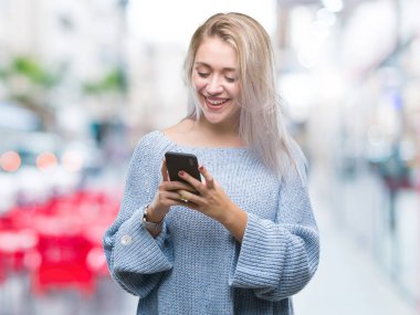 Sarışın genç kadın manifatura izole arka plan üzerinde duran ve diş gösterilen kendine güvenen bir gülümsemeyle gülümseyerek mutlu bir yüz ile Smartphone kullanarak ileti gönderme