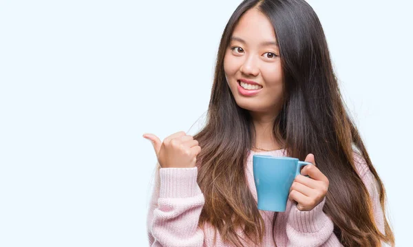 年轻的亚洲妇女喝咖啡在孤立的背景下指向和显示与拇指到一边 快乐的脸微笑 — 图库照片