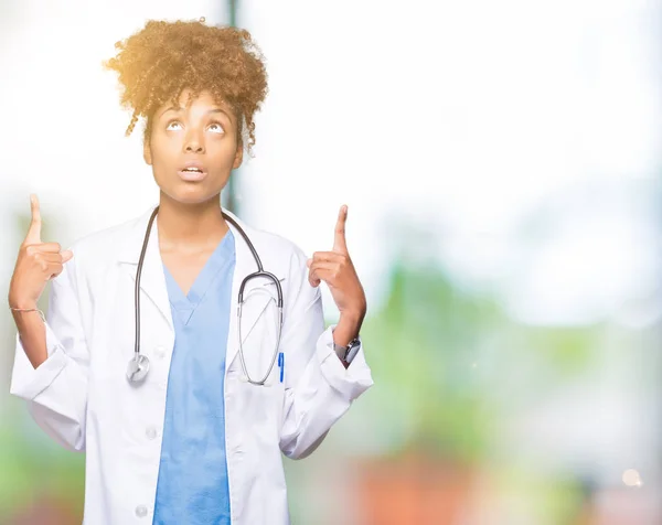 年轻的非洲裔美国医生妇女在孤立的背景惊讶和惊讶地抬起头来 用手指和举起的武器指指点点 — 图库照片