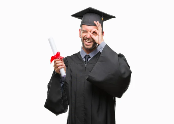 指の間から見て目に手で サインをして笑っている幸せそうな顔で孤立の背景の上の学位を保持若いハンサムな卒業の男 — ストック写真