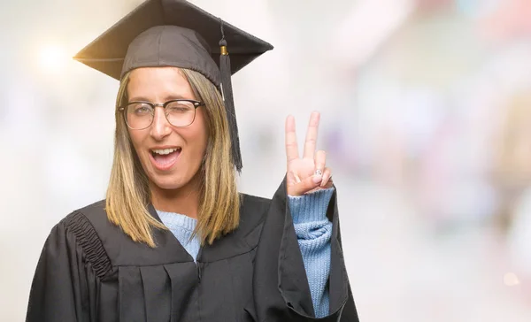着た若い美しい女性は 勝利のサインを行うカメラでウィンクしている幸せそうな顔を浮かべて隔離された背景に制服を卒業しました — ストック写真