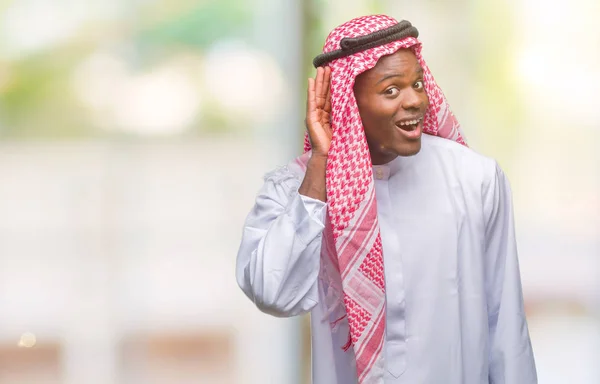 Young arabic african man wearing traditional keffiyeh over isolated background smiling with hand over ear listening an hearing to rumor or gossip. Deafness concept.