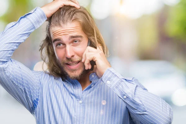 Young handsome man with long hair over isolated background talking on the phone stressed with hand on head, shocked with shame and surprise face, angry and frustrated. Fear and upset for mistake.