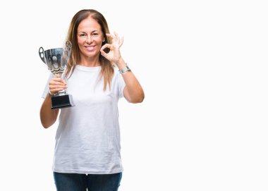 Orta yaş İspanyol kazanan kadın kupa parmak, mükemmel sembolü ile ok işareti yapıyor izole arka plan üzerinde tutarak Ödülü kutluyor