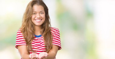 Giyen genç güzel esmer kadın çizgili tişört üzerinde gülümseyen izole arka plan el avuç içi ile birlikte alma veya jest vererek. Ayrı tutma ve koruma