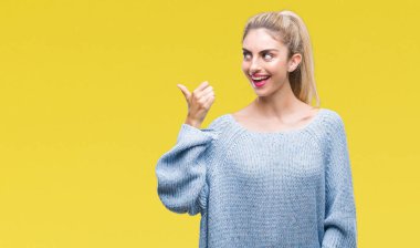 Seyir ve başparmak yanında yukarıyı mutlu yüz ile gülümseyen izole arka plan üzerinde kış kazak giyen genç güzel sarışın ve mavi gözlü kadın.