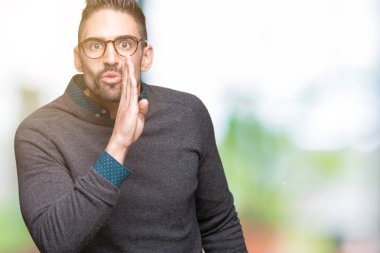 Genç yakışıklı adam gözlük izole arka plan üzerinde kötü amaçlı konuşma konuşma fısıldayan gizli söylenti söylüyor dudaklarından el