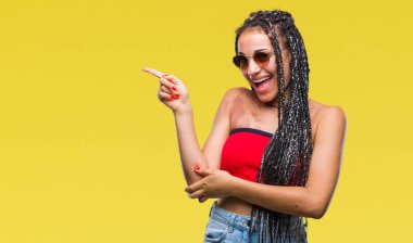 Kameraya bakarak yan el ve parmak ile işaret yüzünde kocaman bir gülümseme ile izole arka plan üzerinde güneş gözlüğü takıyor Doğum işareti ile genç örgülü saç Afrikalı-Amerikalı.