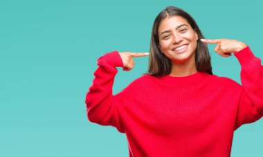 Kendinden emin gösterilen ve işaret parmakları diş ve ağız ile gülümseyen izole arka plan üzerinde kış kazak giyen genç güzel Arap kadın. Sağlık kavramı.