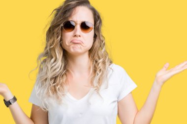 Güzel genç sarışın kadın üzerinde güneş gözlüğü takıyor arka plan clueless ve şaşkın bir ifadeyle kollar ve eller kaldırdı izole. Şüphe kavramı.