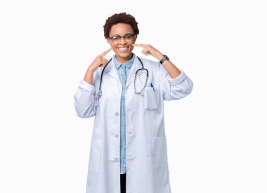Genç Afro-Amerikan doktor kadın kendinden emin gösterilen ve işaret parmakları diş ve ağız ile gülümseyen izole arka plan üzerinde tıbbi bir palto giyiyor. Sağlık kavramı.